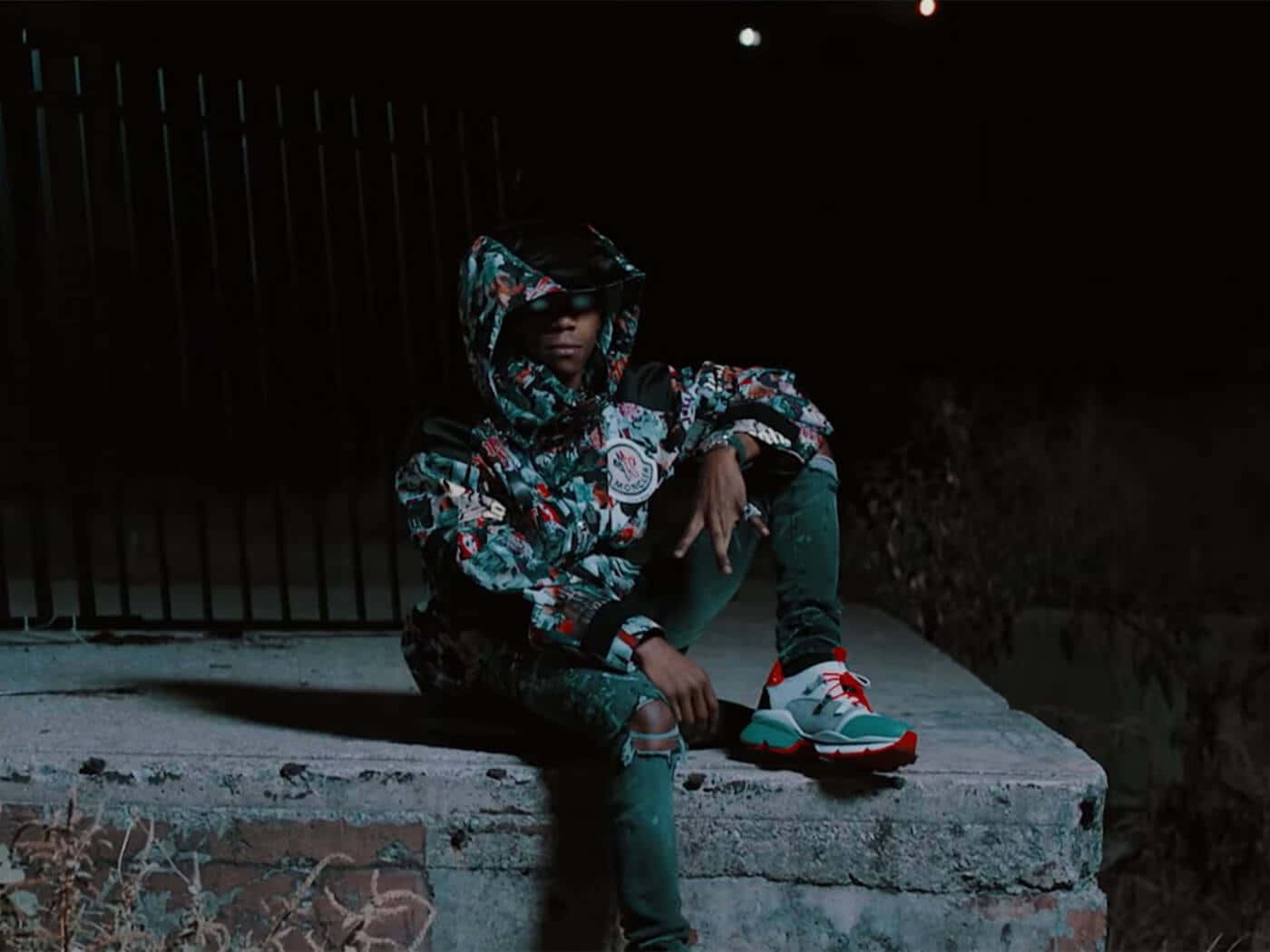 A Man Sitting On A Wall At Night Wearing A Hoodie And Sneakers Wallpaper