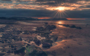 A Beach With Rocks And A Sun Setting Behind It Wallpaper