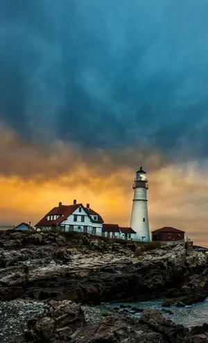 A Lighthouse Sits On A Rocky Shore With A Cloudy Sky Wallpaper