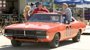 A Man And Woman Pose In Front Of An Orange Muscle Car Wallpaper