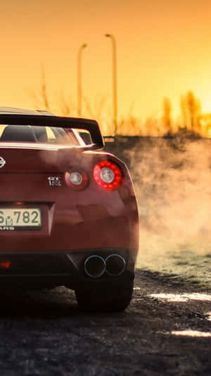 A Red Sports Car Is Driving Down The Road At Sunset Wallpaper