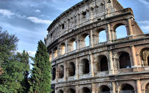 Caption: Majestic Colosseum Underneath A Cloudy Sky Wallpaper