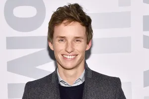 Eddie Redmayne Smiling In A Black Suit Against A Maroon Background Wallpaper